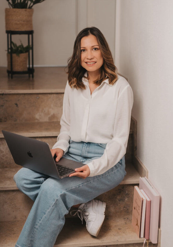 Mary Webdesigerin in Nidda, sitzt mit einem laptop auf dem Schoß auf einer Treppe und lächelt in die Kamera